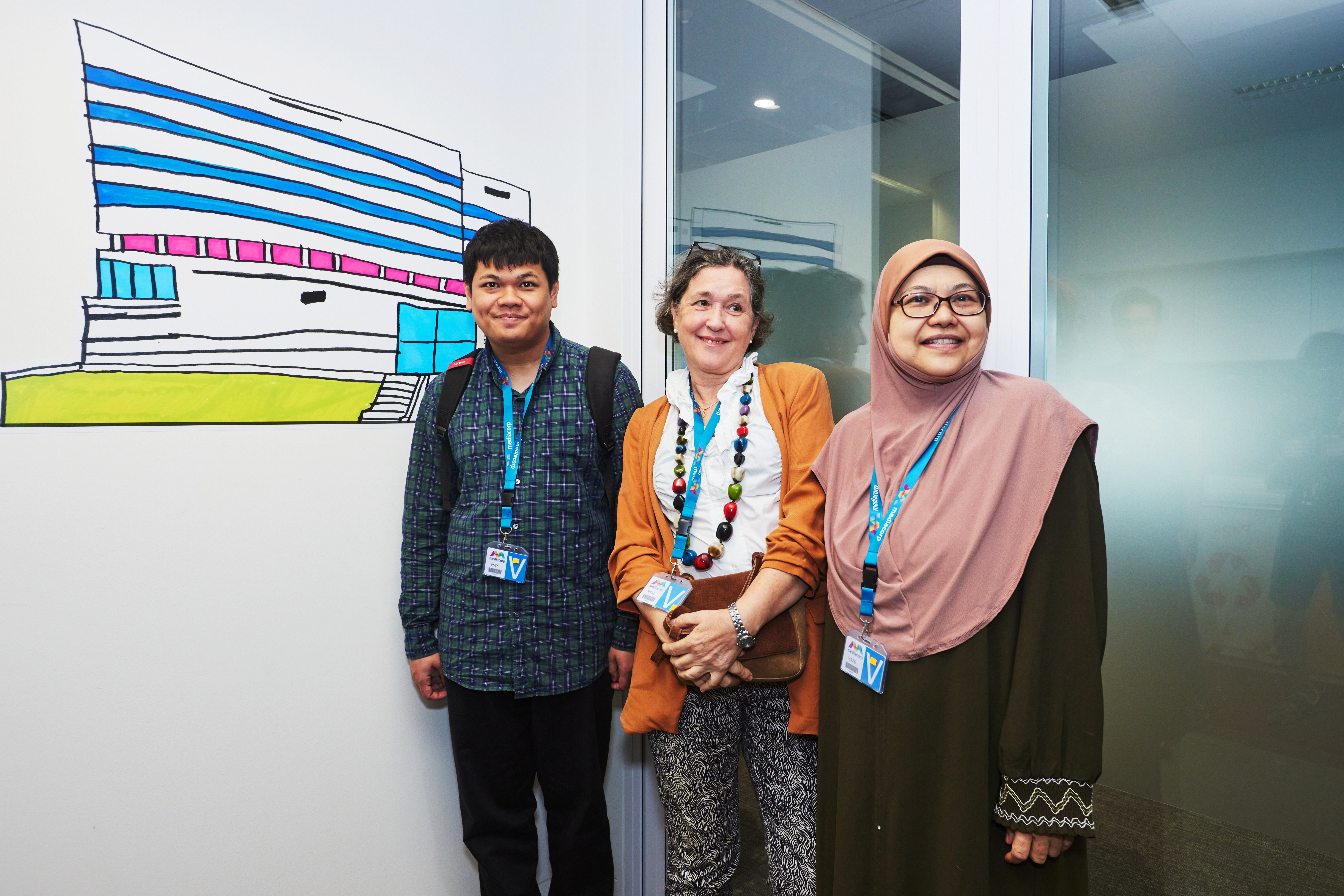 Muhammad Fadhil - Artist with autism from Metta Welfare Association, stands with his mother and Dr Esther Joosa in front of his sticker mural that features Mediacorp Campus building