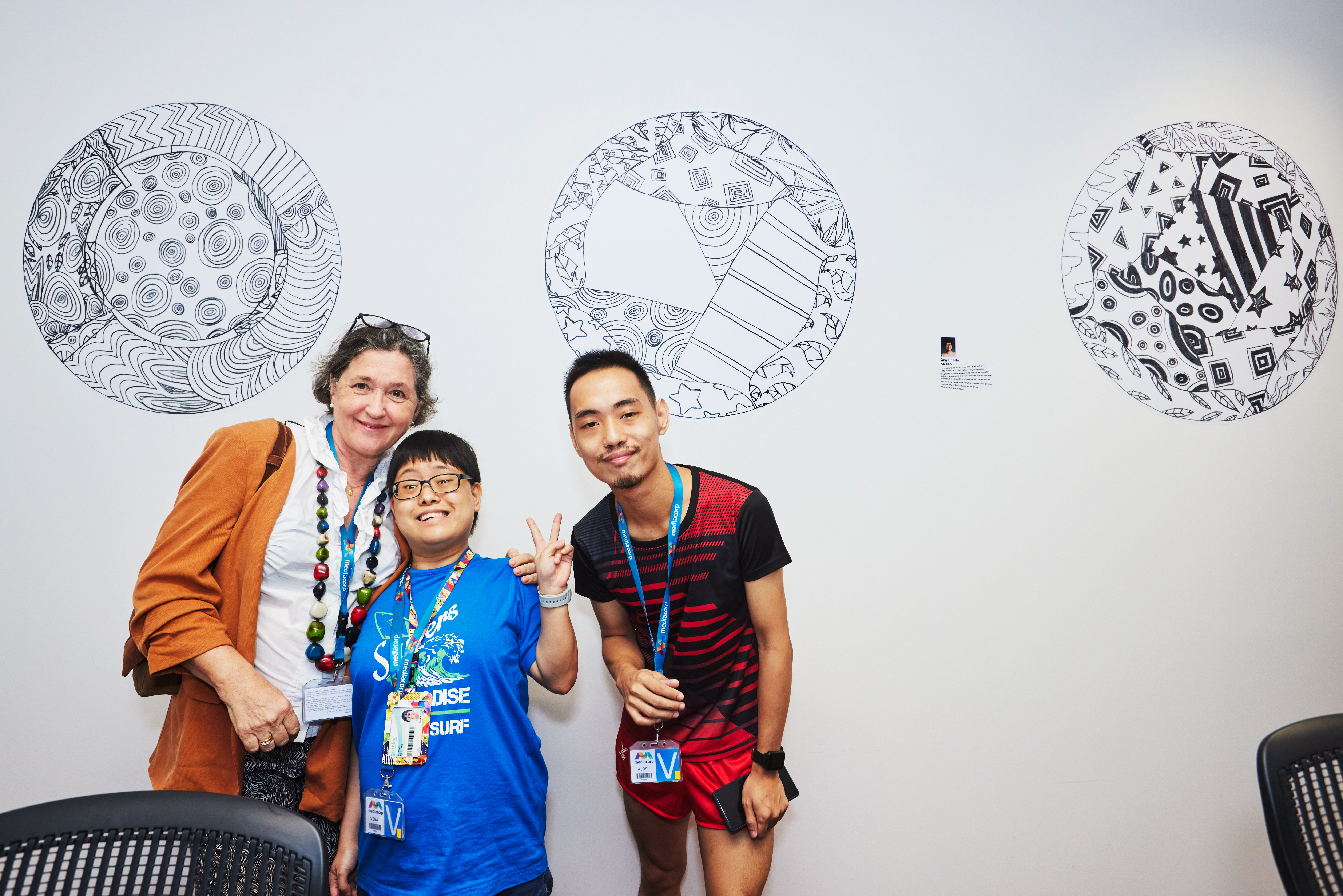 Ong Xin Min - Artist from MINDS, poses with hand in 2 finger peace sign with her brother and Dr Esther in front of circular wall sticker murals of intricate lines and patterns 