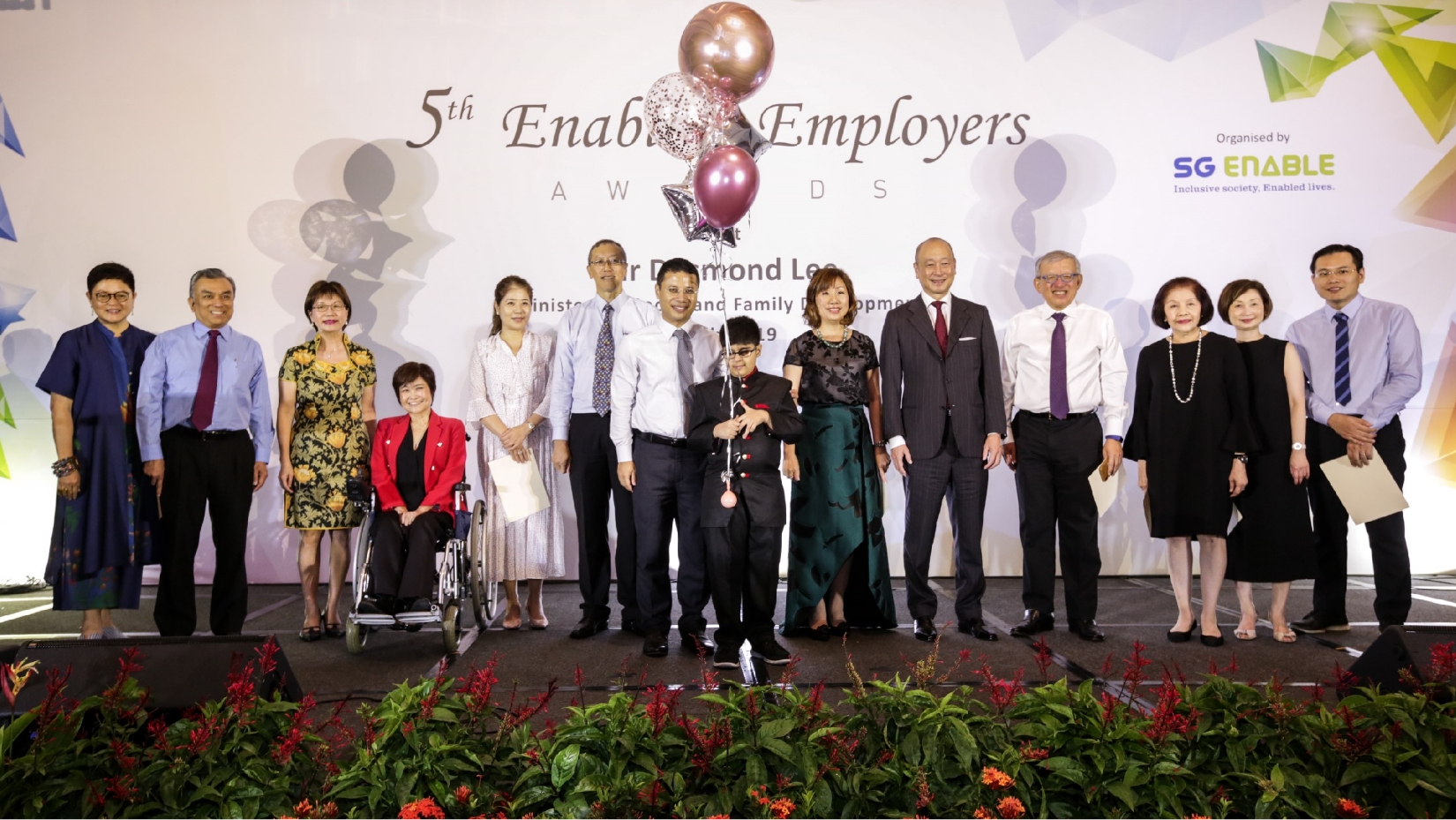 About a dozen VIPs standing on the stage at the 5th Enabling Employers Awards. In the centre, a boy holds balloons.